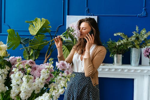 Woman in Brown Long Sleeve Shirt Smelling Pink Flowers while Talking on the Phone