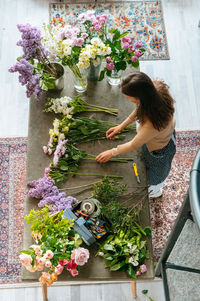 Kvetinár s dlhými vlasmi v pracovnom oblečení opatrne viaže kyticu z čerstvých kvetov v svetlom floristickom ateliéri