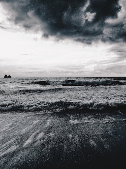 Black and white of wavy ocean near sandy coast under cloudy sky in stormy weather