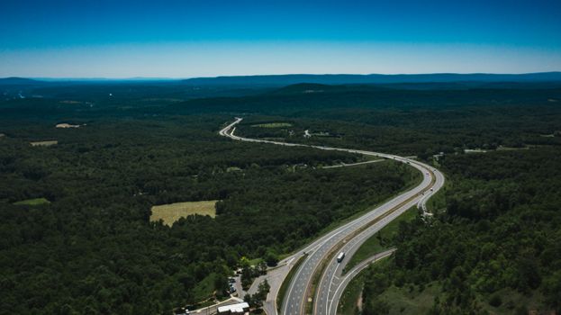 Drone view of asphalt road with driving vehicles between lush green trees in woods under colorful sky