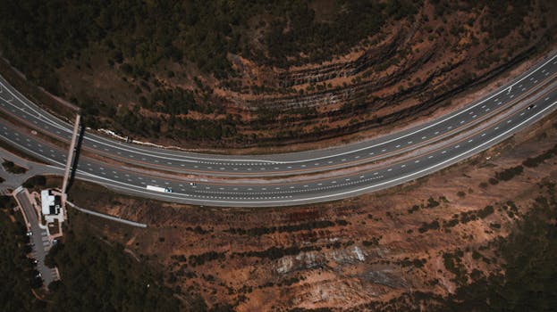 Overhead view of wavy road with marking lines and vehicles between brown terrain with trees and barrier