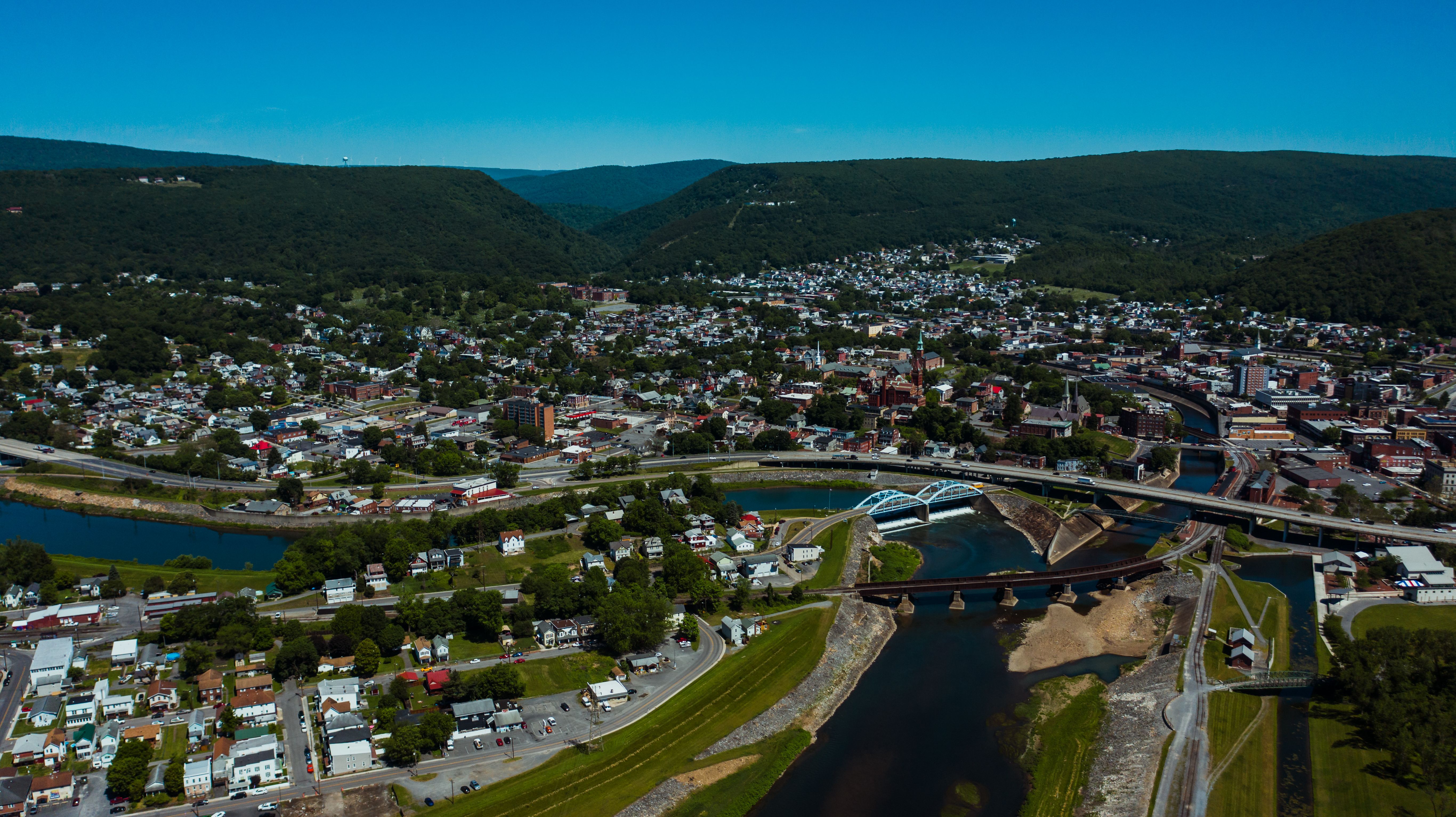 Aerial view of wavy roadway and bridges over canal between dwelling buildings near lawns with trees in mounts under blue sky
