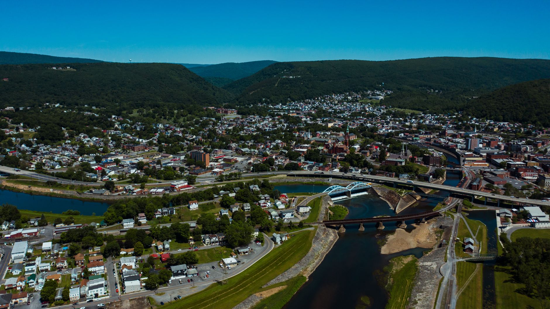 Aerial view of wavy roadway and bridges over canal between dwelling buildings near lawns with trees in mounts under blue sky