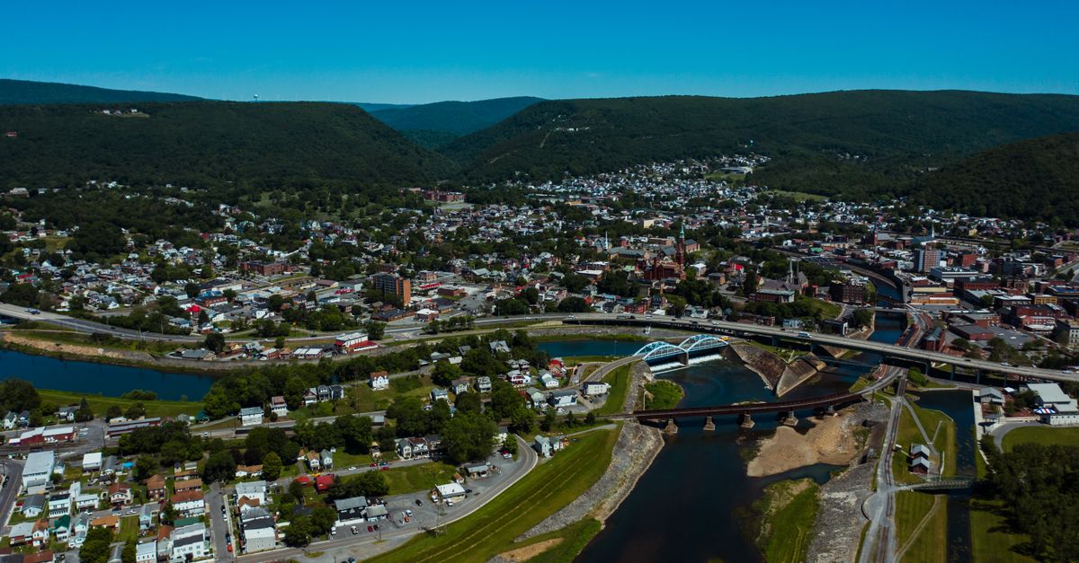 Aerial view of wavy roadway and bridges over canal between dwelling buildings near lawns with trees in mounts under blue sky