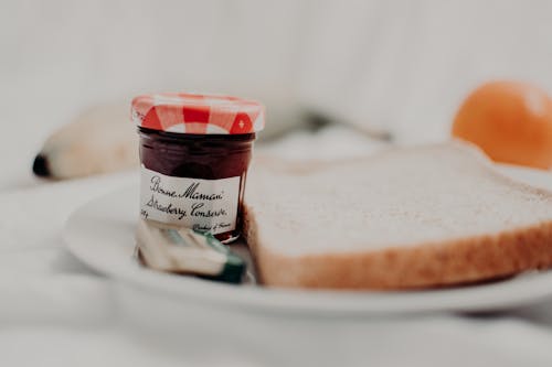Tasty marmalade in glass jar with written title near bread loaf for breakfast on plate