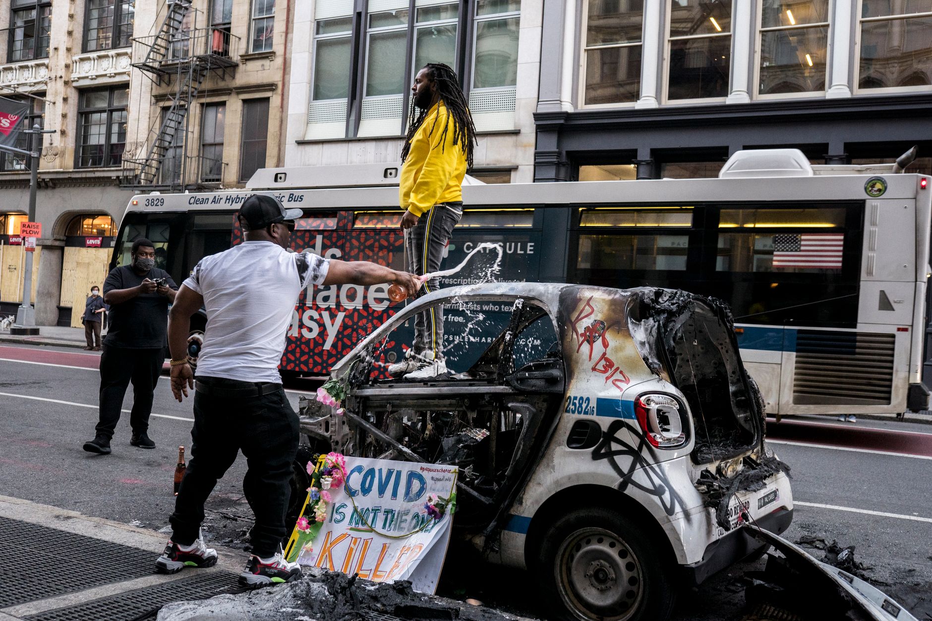 Protesters in urban setting with burnt car and COVID sign.
