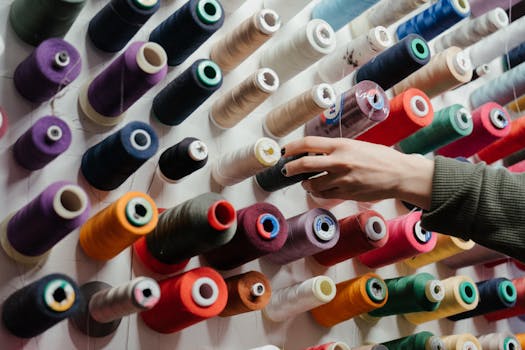 Hand reaching for a spool of thread from a vibrant wall display of organized sewing materials.
