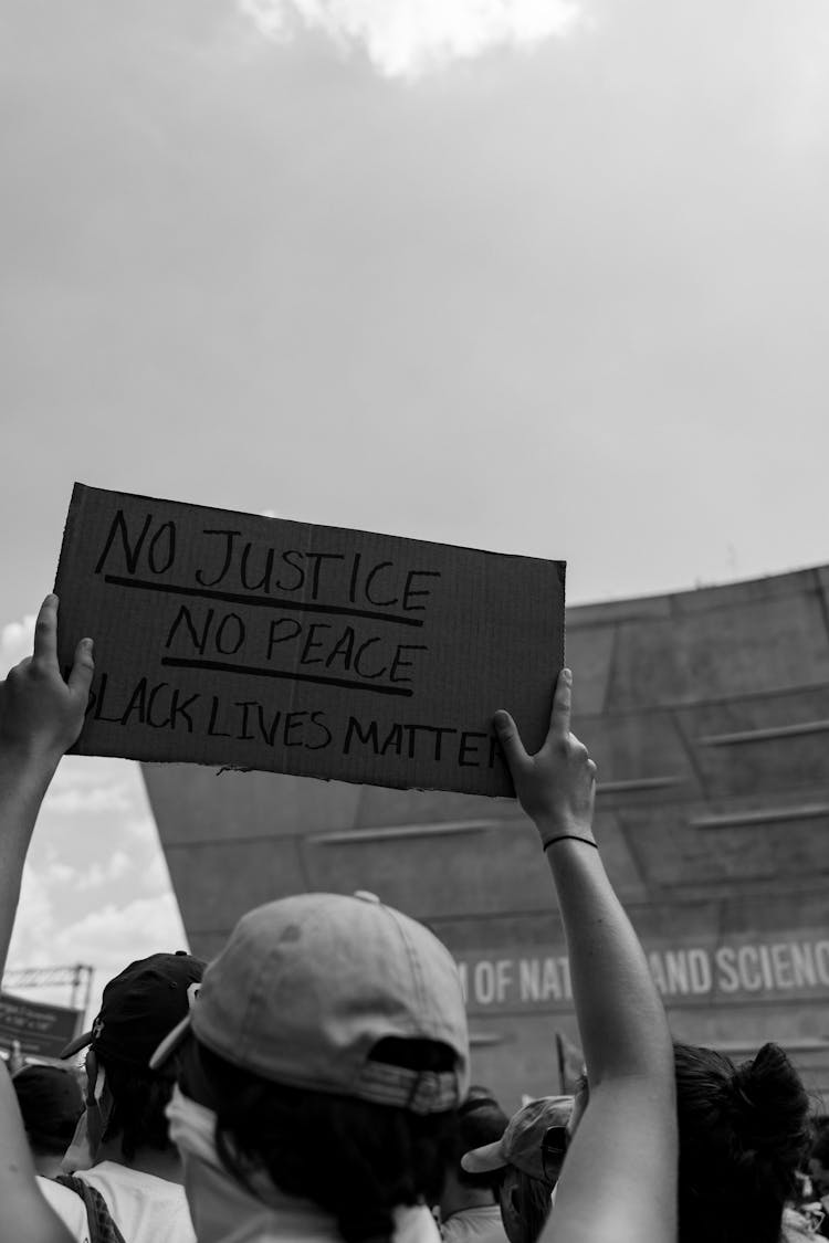 Grayscale Photo Of Person Wearing A Cap Holding A Cardboard With Message 