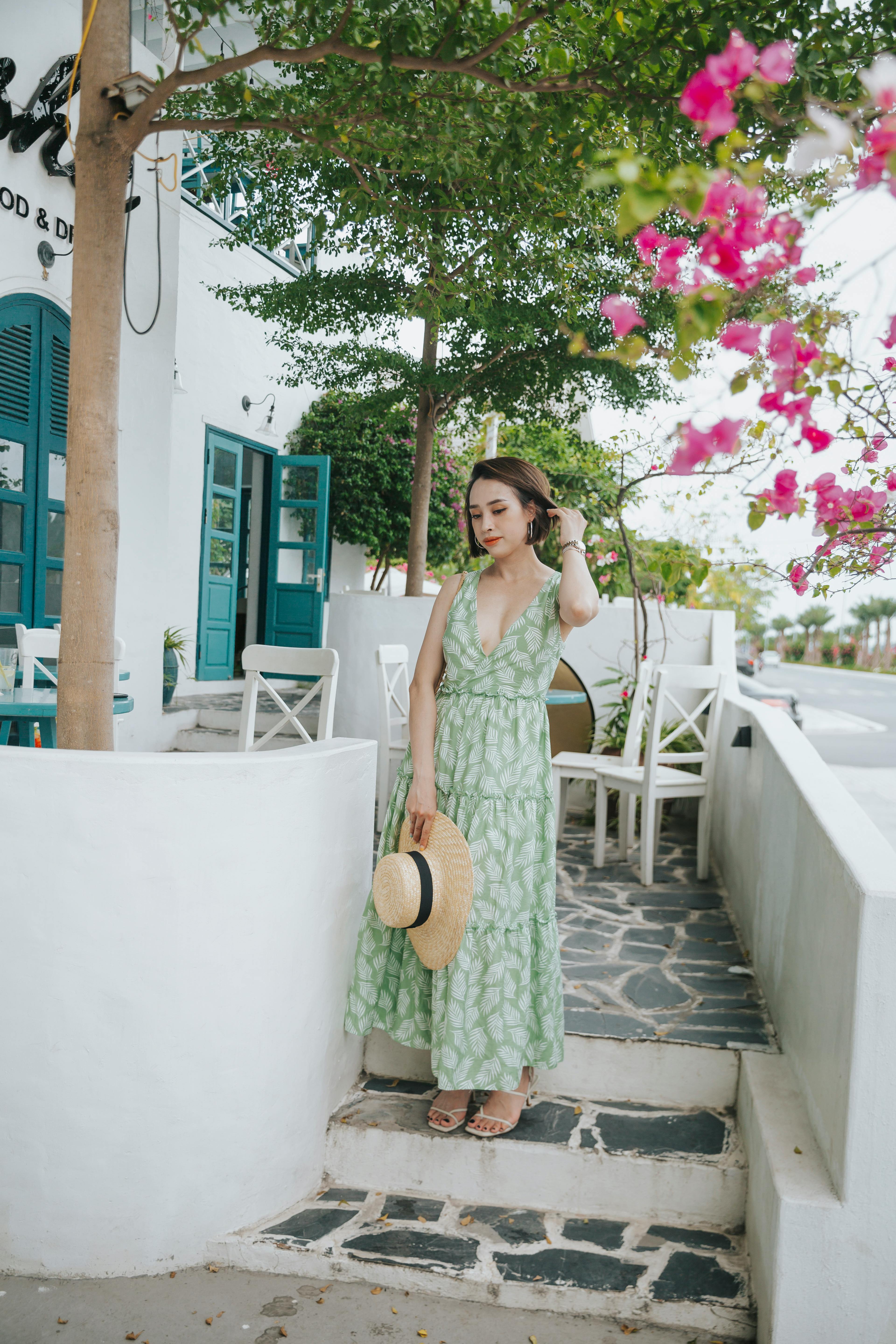 Young pondering ethnic lady in trendy apparel with straw hat touching hair while looking down on staircase
