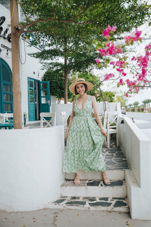Free Full body of cheerful female in summer dress and hat standing on steps of street cafe and looking at camera with toothy smile Stock Photo