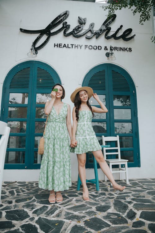 Full body of positive female friends in stylish summer dresses holding hands standing on terrace of building on city street