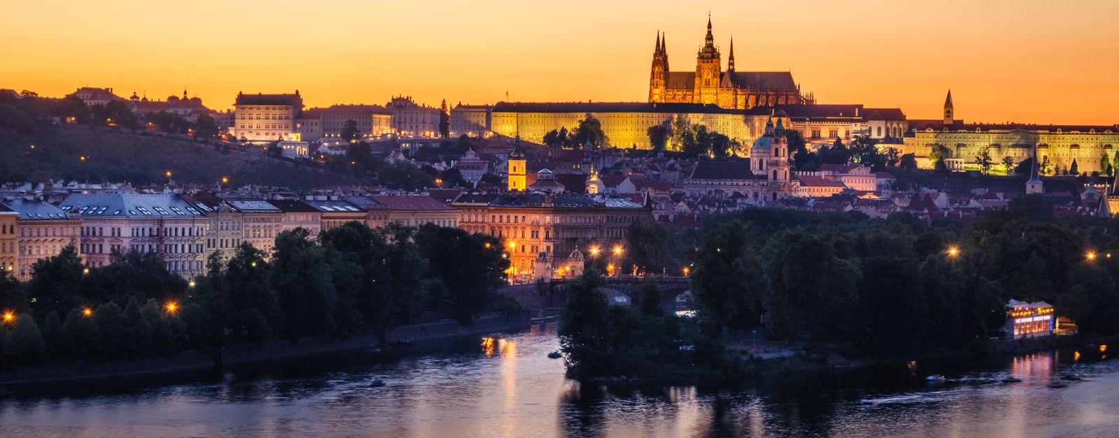 Scenic view of Prague Castle glowing at sunset with the Vltava River in the foreground.