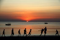 在日落时分，海滩边玩耍的人的剪影映入眼帘，而他们身后则是一艘小船