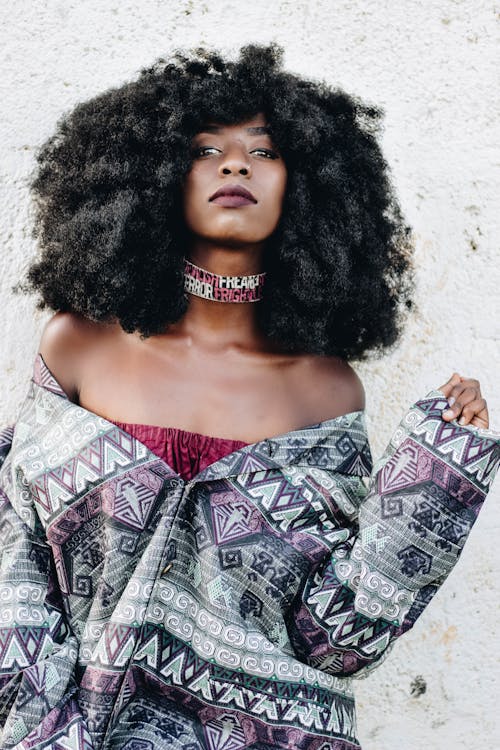 Young trendy African American female in modern clothes and necklace looking at camera near rough wall