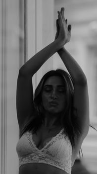 Black and white of young provocative female in lace lingerie standing with raised arms and pouting lips near window in apartment while looking away