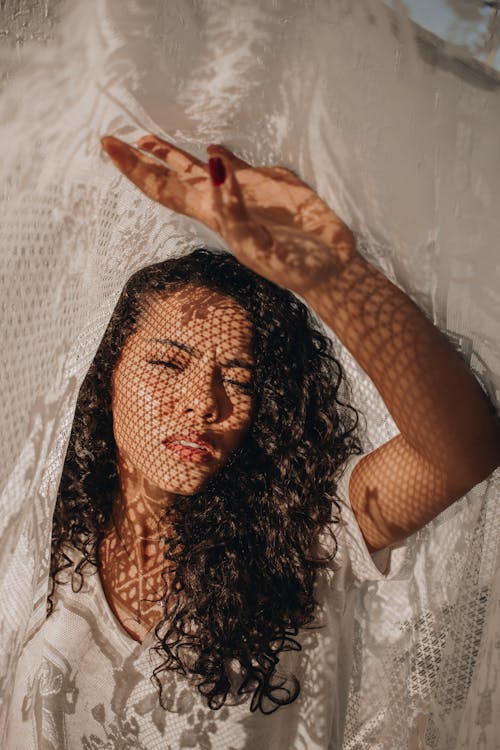 Young ignorant trendy female with curly hair standing with raised arm with shadow of curtain on face in sunlight and looking at camera