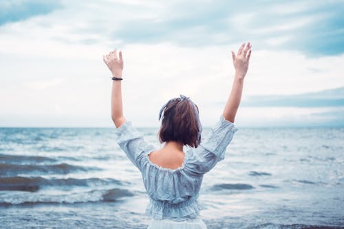 Serene faceless woman standing with arms raised near waving sea