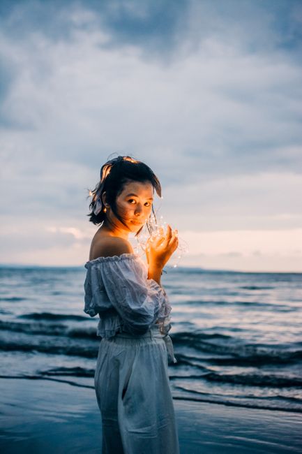 Side view of stylish young Asian female in summer dress with bare shoulders standing on evening seashore with glowing garland in hands