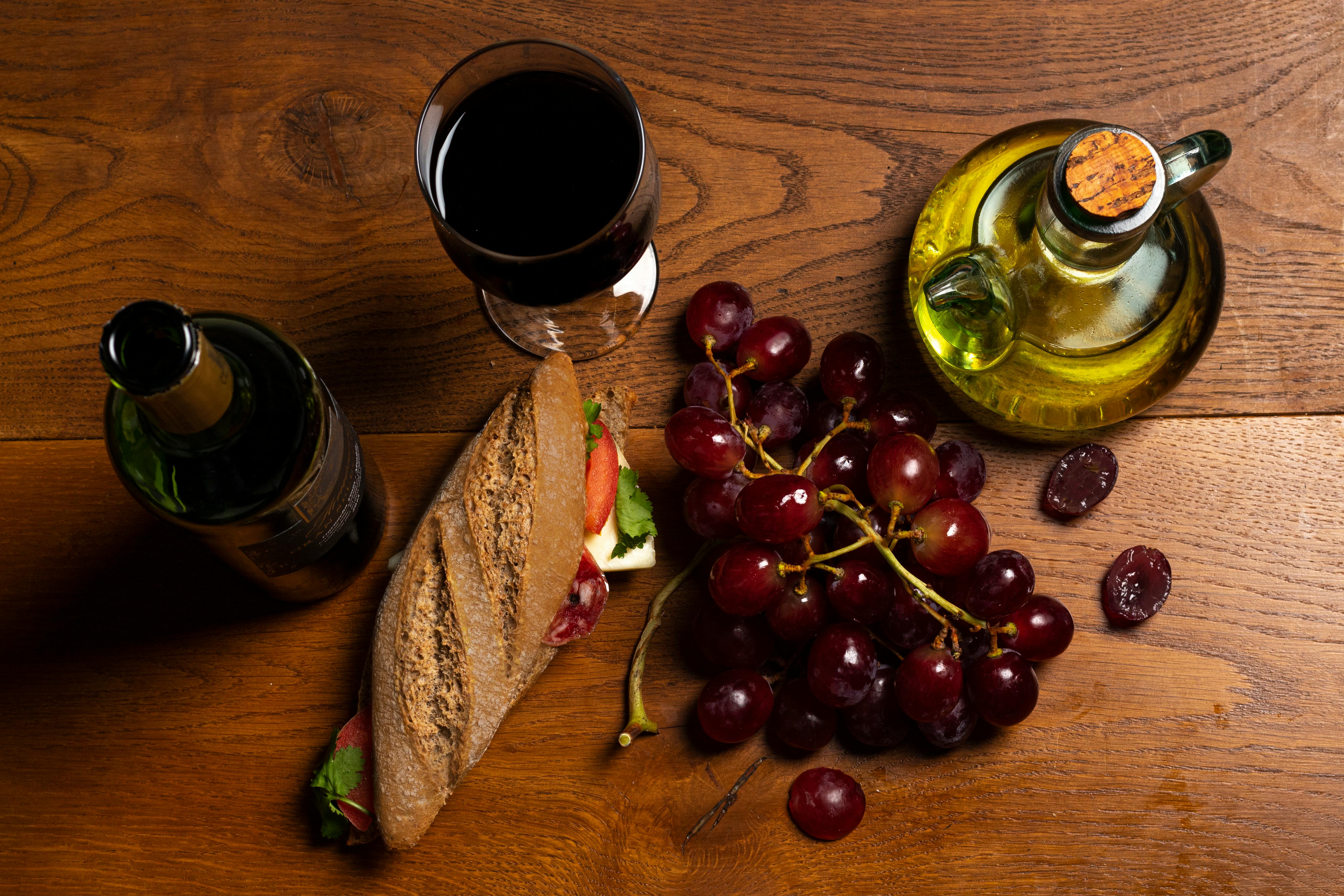 Top view of a rustic meal with wine, baguette, grapes, and olive oil on a wooden table.