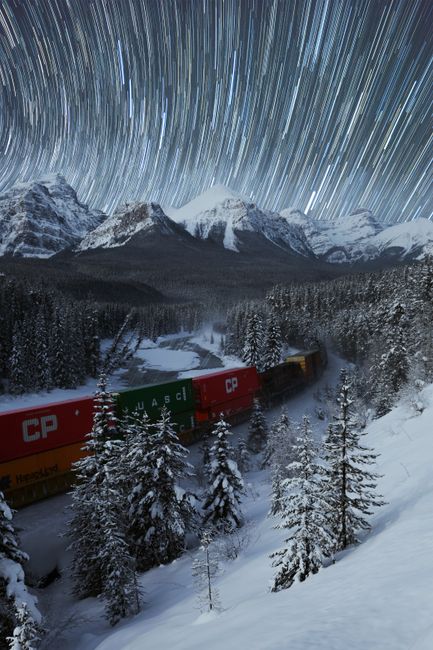From above of train driving on snowy mountainous terrain near coniferous trees against starry night sky