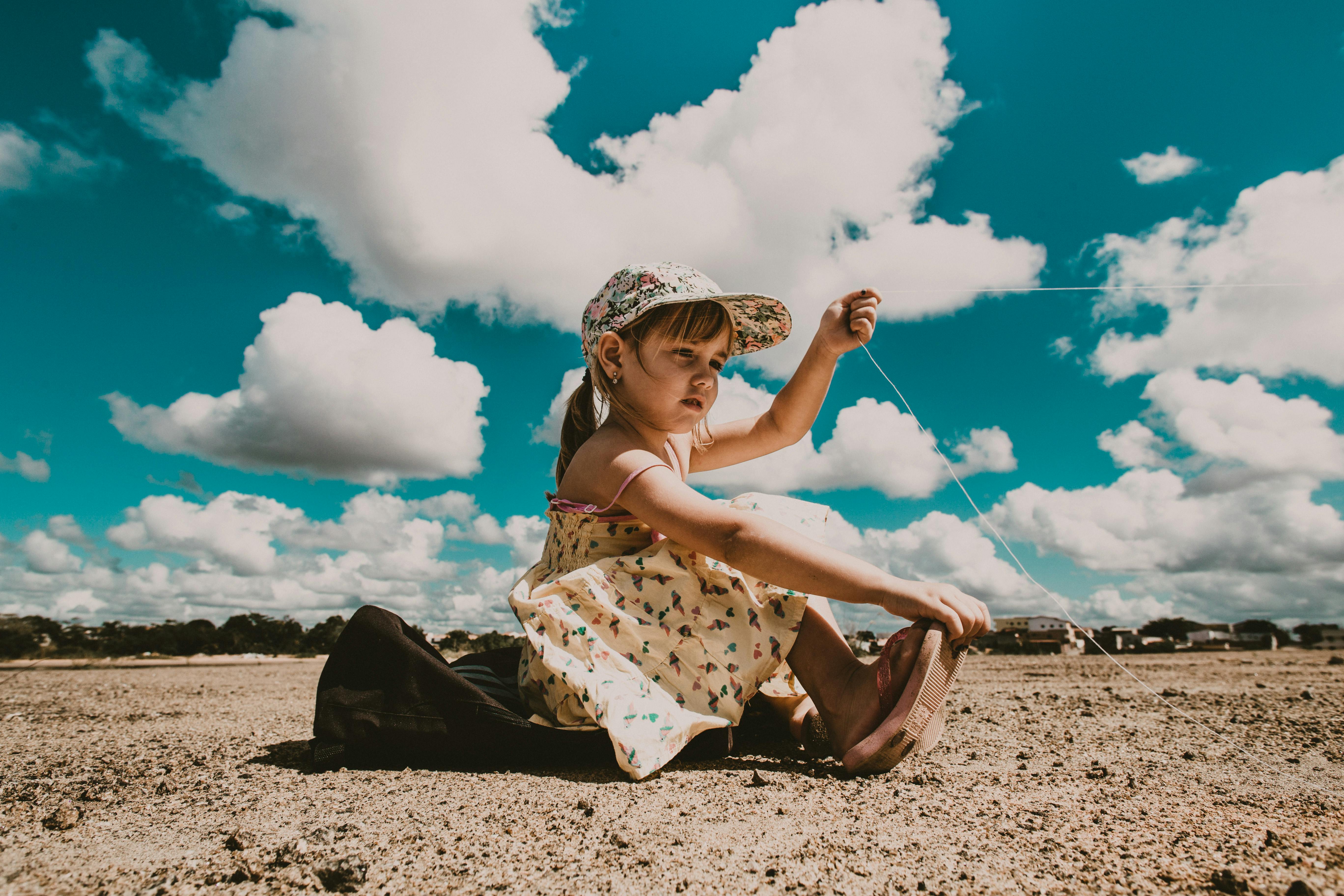 Side view of adorable little girl in dress and cap sitting on ground and holding kite thread against cloudy blue sky