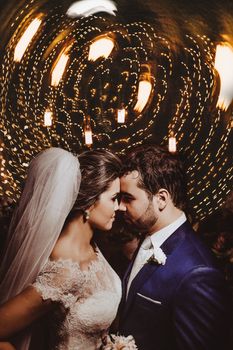 Side view of young bearded groom in elegant suit and gorgeous bride in lace white dress and veil touching foreheads and looking at each other against glowing garlands