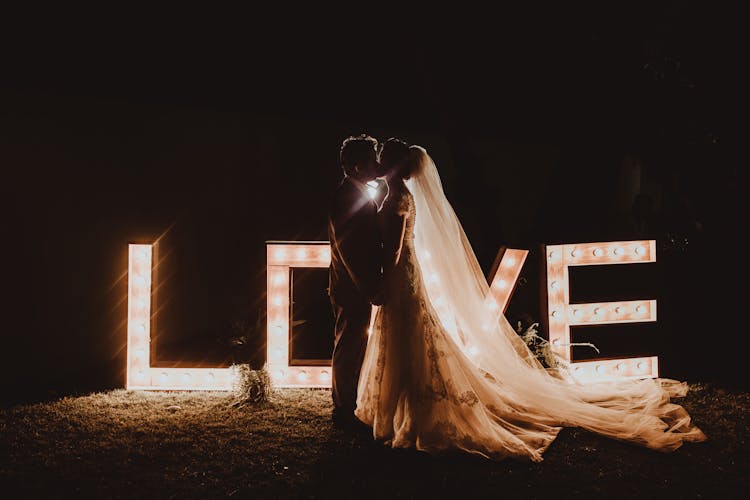 Anonymous Newlywed Couple Kissing At Night Near Illuminated Love Light Bulb Letter