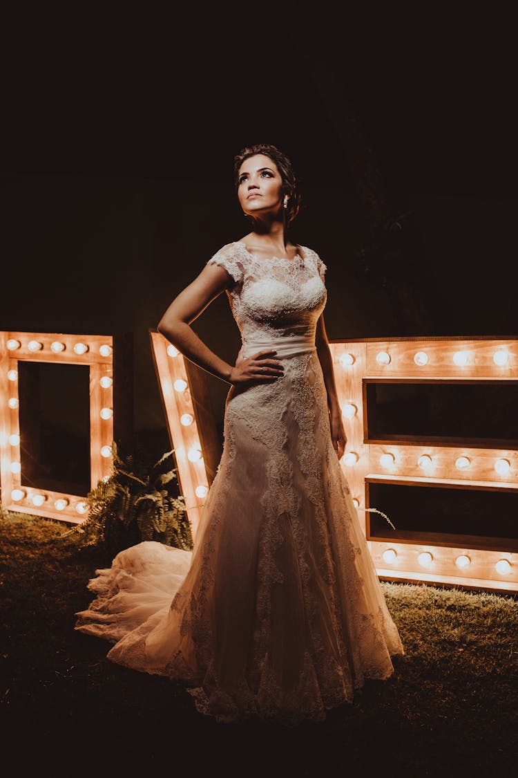 Graceful Young Bride Standing In Dark Garden Near Light Bulb Letter