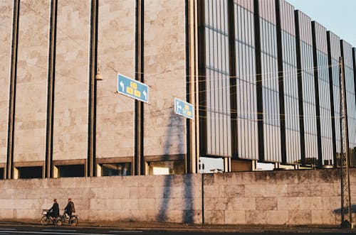 Free Anonymous people riding bicycles on asphalt road near modern building with geometric facade in sunny morning Stock Photo