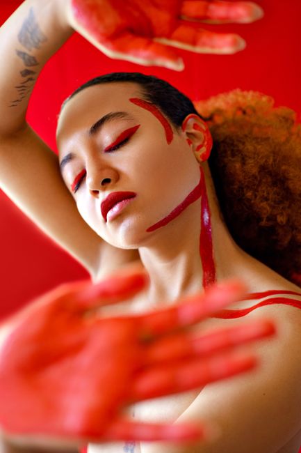 Naked tattooed young ethnic female with closed eyes standing against red background with painted face and hands