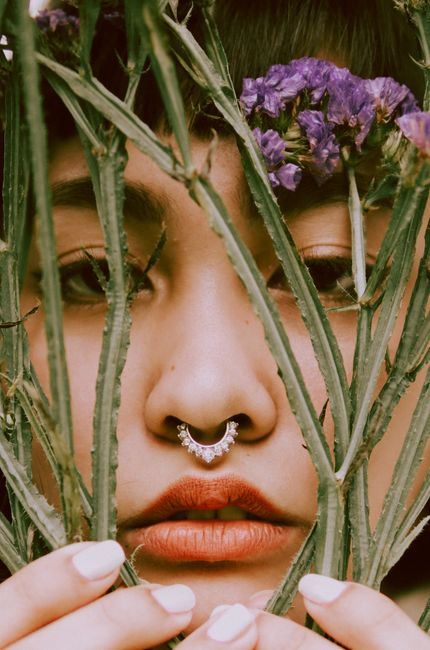 Artistic close-up portrait of a woman with nose piercing framed by cactus stems and purple flowers.
