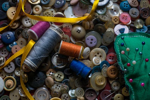 Overhead of coils of thread and round multi colored buttons composing with glossy narrow yellow ribbon and needles in pin cushion