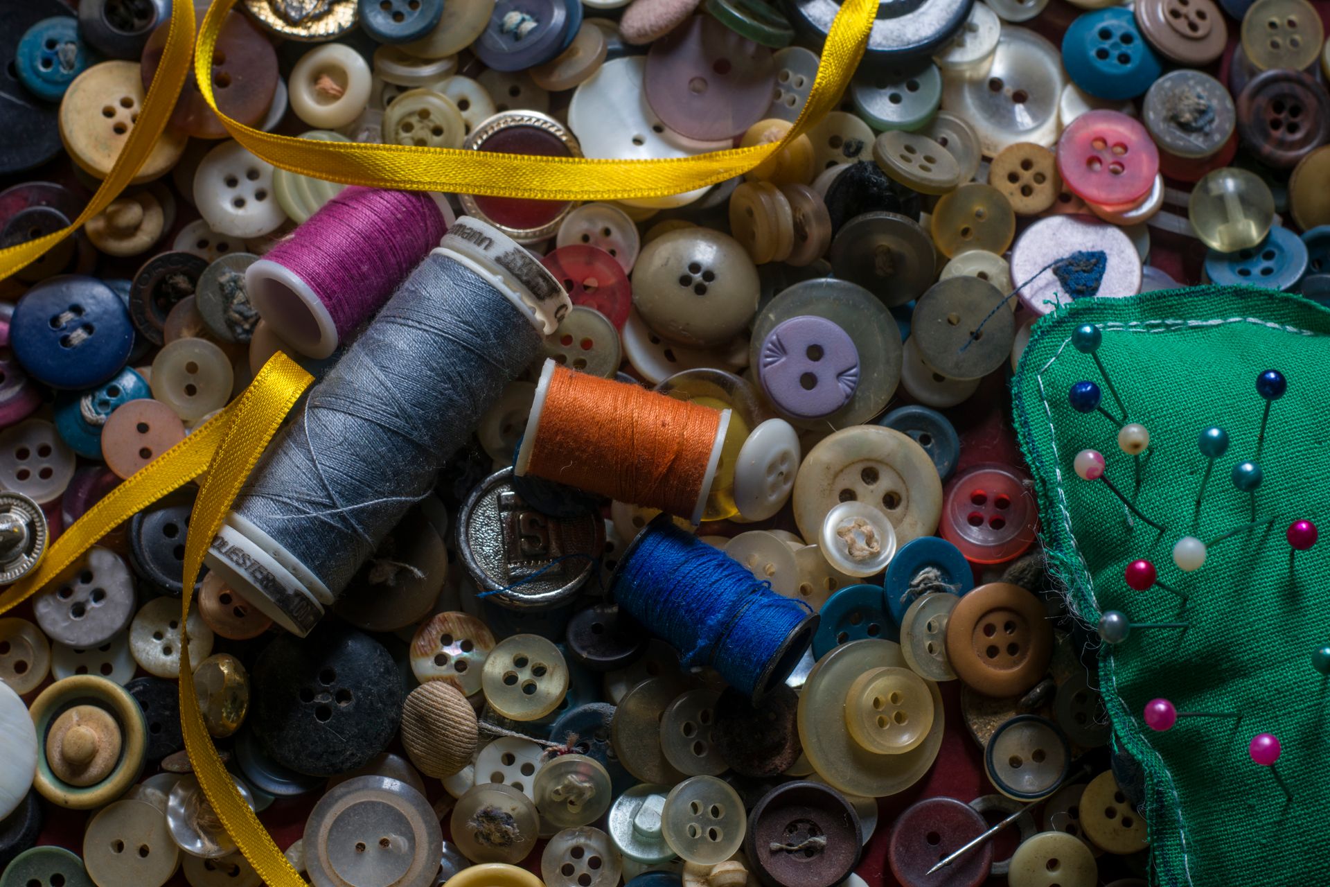 Overhead of coils of thread and round multi colored buttons composing with glossy narrow yellow ribbon and needles in pin cushion