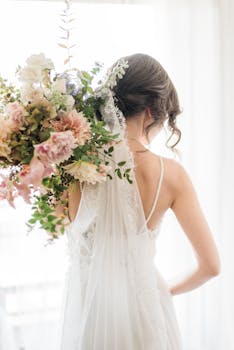A bride elegantly poses with a floral bouquet in a white wedding dress indoors.