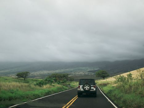 A jeep travels down a winding rural road surrounded by lush scenery under a cloudy sky.
