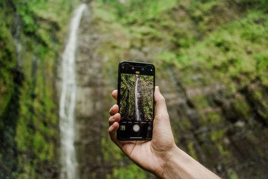 Close-up of a hand holding a smartphone taking a photo of a waterfall.
