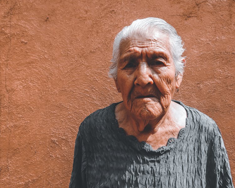 Old Woman Near Beige Wall In Sunlight