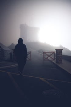 A silhouette of a person walking uphill in a foggy, backlit landscape, creating an eerie atmosphere.
