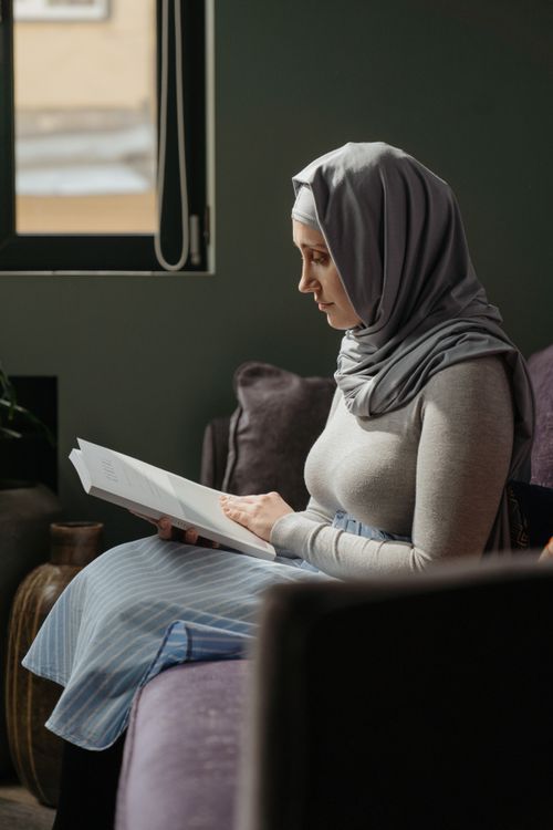 Free A woman wearing a hijab sits on a sofa reading a book in a warm, inviting living room setting. Stock Photo