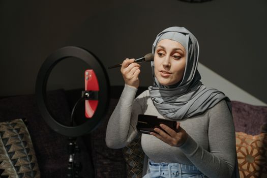 A woman in a hijab applies makeup in a beauty tutorial setting with a ring light.