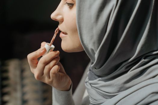 Close-up of a woman in a hijab applying lip gloss indoors, showcasing beauty routine.