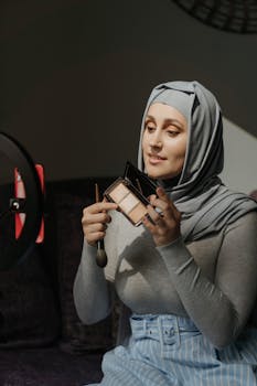 A woman in a hijab demonstrating a makeup tutorial at home using a smartphone.