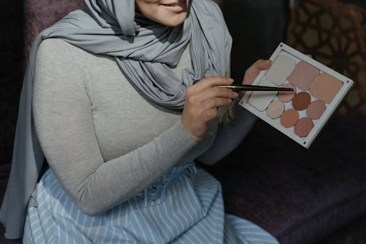 Muslim woman in a hijab holding an eyeshadow palette indoors, showcasing makeup.