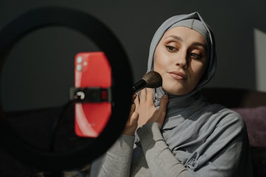 A Muslim woman in a hijab applies makeup for a tutorial indoors.