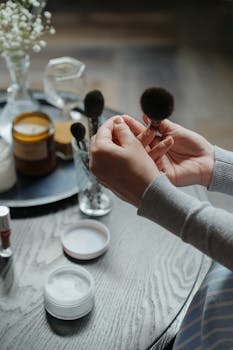 A woman's hands holding a makeup brush over a tabletop with cosmetics in a warm, indoor setting.