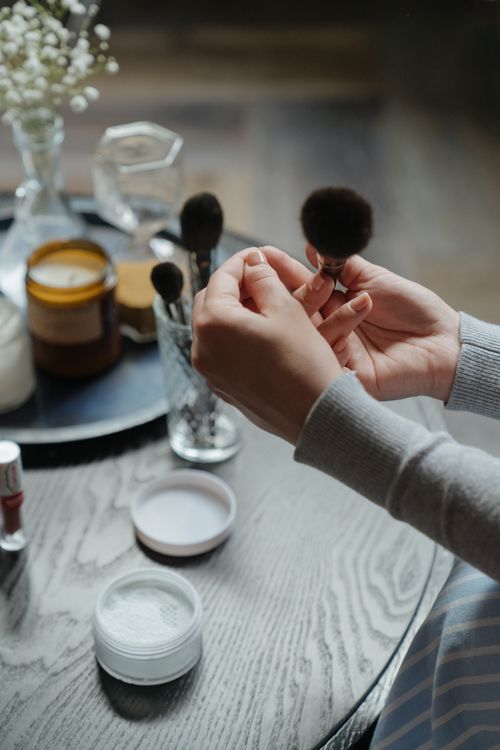 Free A woman's hands holding a makeup brush over a tabletop with cosmetics in a warm, indoor setting. Stock Photo