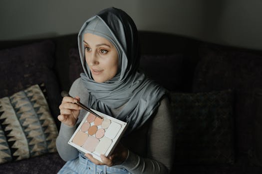 A woman wearing a hijab applies eyeshadow indoors, showcasing beauty and cosmetics.
