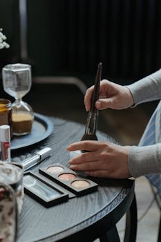 A close-up of a woman's hands applying makeup with brushes and an eyeshadow palette indoors.