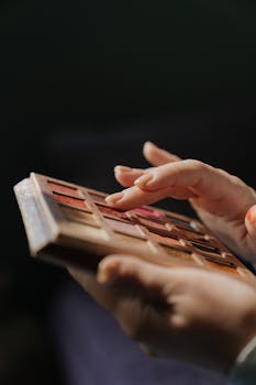 A close-up view of hands applying eyeshadow from a colorful palette indoors.