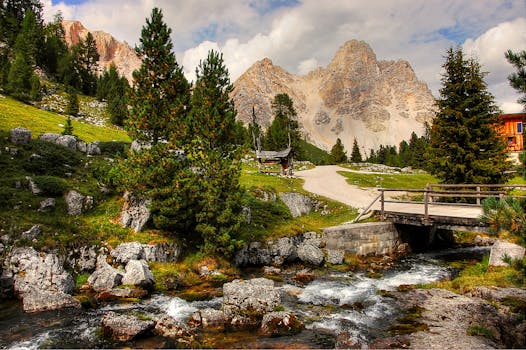 A beautiful mountain landscape with a flowing stream, lush greenery, and a bridge under a cloudy sky.